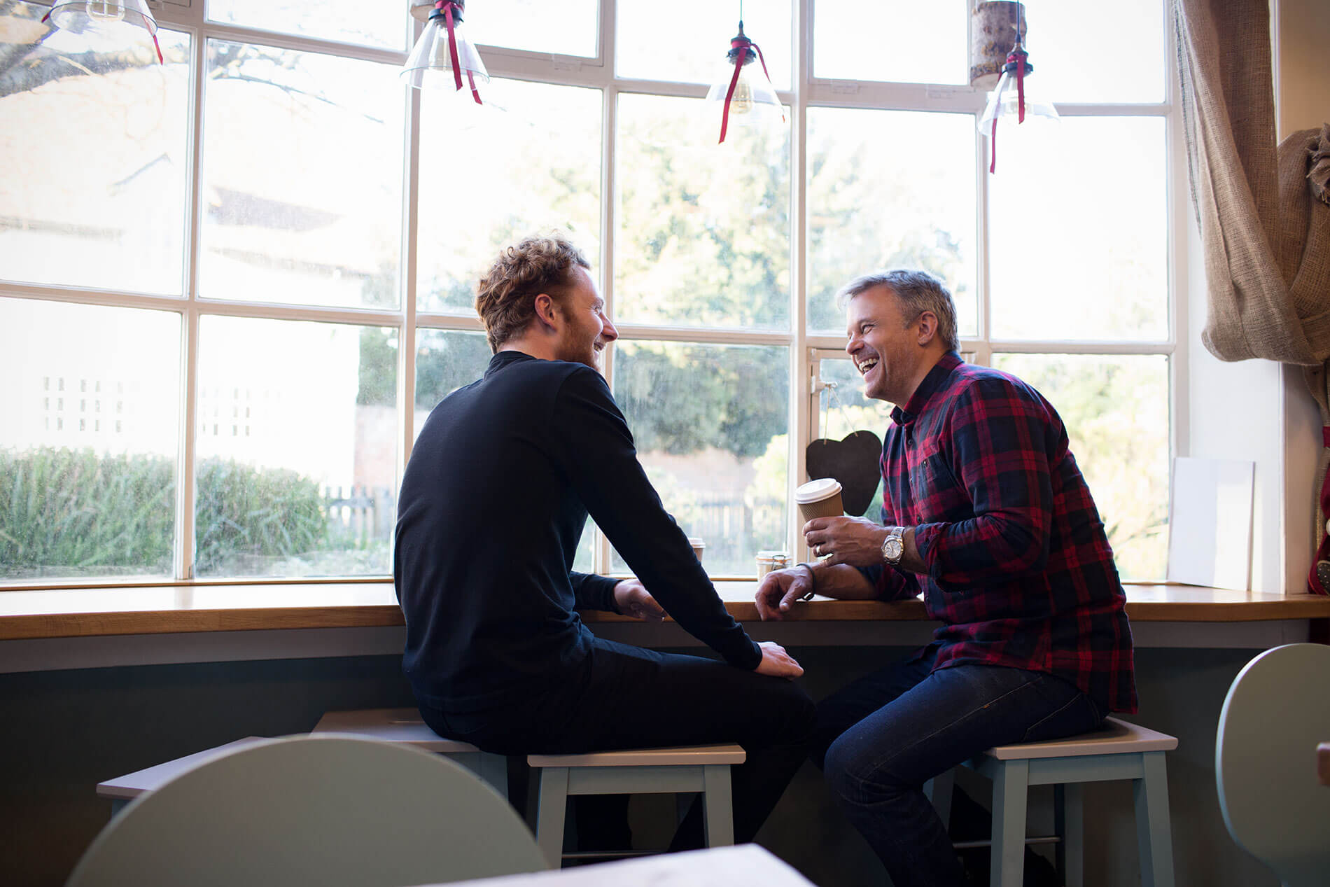 Two men having a conversation and laughing together at a café table by the window.